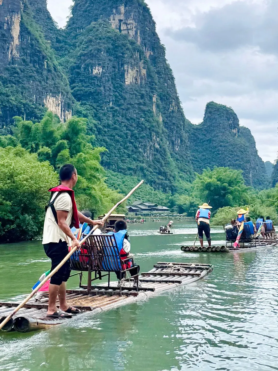 Several bamboo rafts carrying tourists in blue life vests float on the calm green Yulong River. A raft guide stands on the foreground raft, pushing with a long pole. Majestic, tree-covered karst mountains rise steeply in the background under a cloudy sky, with green bamboo forests along the riverbanks.