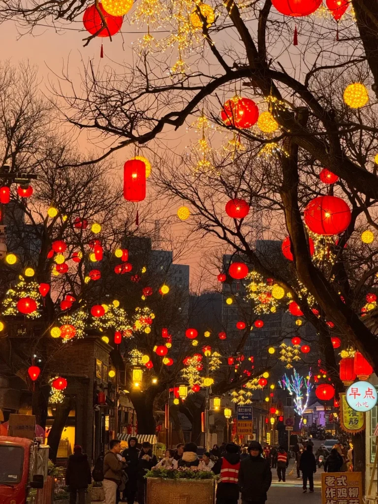 A festive street scene at dusk adorned with numerous red lanterns and yellow starburst fairy lights hanging from the bare branches of trees. People are walking along the bustling street below, which is lined with traditional-style buildings and glowing shop signs. The sky above is a soft orange and pink.