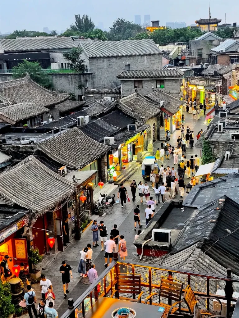 Aerial view of a narrow, lively Beijing Hutong street at evening. Traditional grey-tiled roof buildings with illuminated shop fronts line both sides, bustling with pedestrians. Red lanterns are visible near several entrances. In the foreground, a rooftop patio with tables and chairs overlooks the street, and tall modern buildings are faintly visible on the horizon.