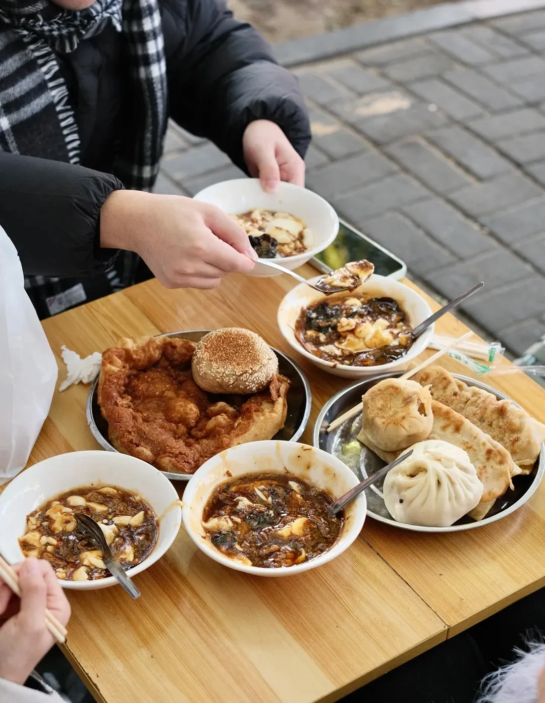 A wooden table laden with various Beijing street food breakfast items, including multiple bowls of savory douhua (tofu pudding) with dark sauce and mushrooms, a large fried flatbread, a sesame seed bun, a steamed baozi, and pan-fried buns. Two people are visible, one scooping douhua and another holding chopsticks, with a paved ground in the background.