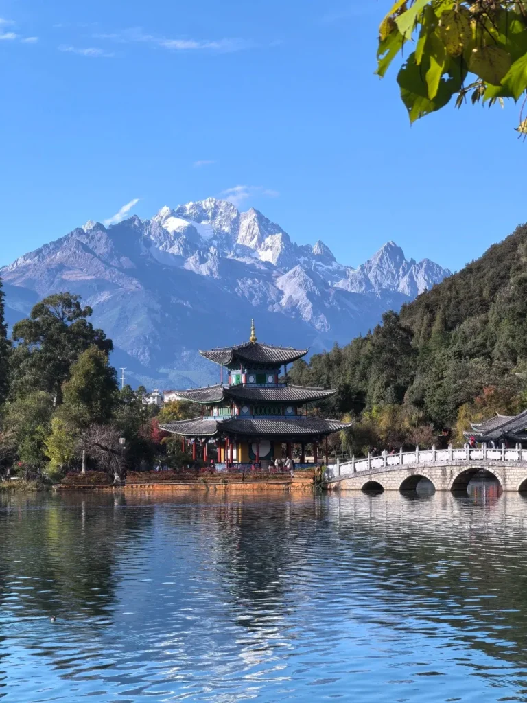 A breathtaking view of the multi-tiered Deyue Pavilion, a traditional Naxi architecture pagoda, located in Black Dragon Pool (Heilongtan Park) in Lijiang, Yunnan, China. The pavilion's vibrant colors and tiered roofs are perfectly reflected in the calm blue water. Majestic, snow-capped peaks of Jade Dragon Snow Mountain rise in the background under a clear sky, with a graceful stone arched bridge visible over the lake to the right.