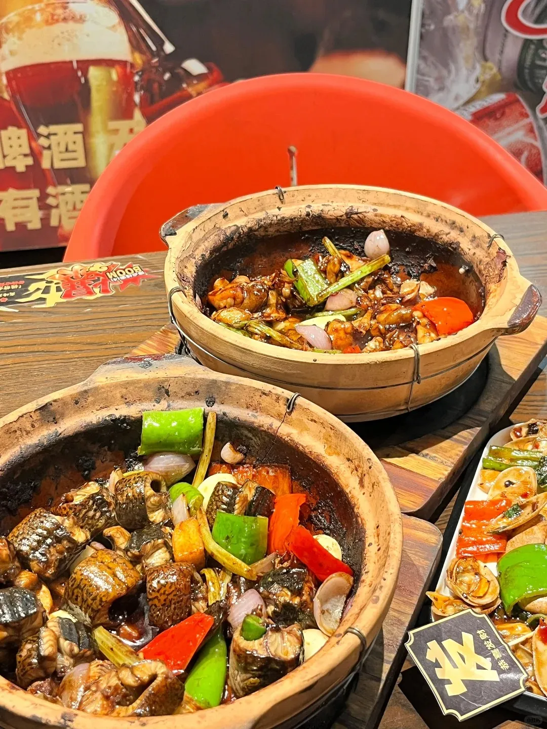 Two traditional Cantonese clay pots, steaming hot, filled with generous portions of braised freshwater eel or fish segments, vibrant green and red bell peppers, crisp white onions, and green onions, all simmered in a dark, savory sauce. The clay pots show rustic, charred edges from cooking. In the background, another dish with clams and vegetables is visible on a rectangular plate, alongside a blurred restaurant poster featuring beer and Chinese characters.