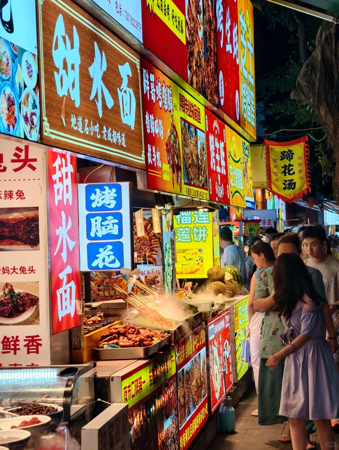 A bustling night food street in Chengdu, China, with numerous brightly lit stalls and crowds of people. Vendors are actively grilling various skewers and other foods like chicken wings, with steam rising from the grills. Large, colorful signs in Chinese advertise dishes such as Sweet Water Noodles (甜水面), Brain Flower (脑花), and Pig Trotter Soup (蹄花汤). Fresh durians are also visible on one of the food stands.