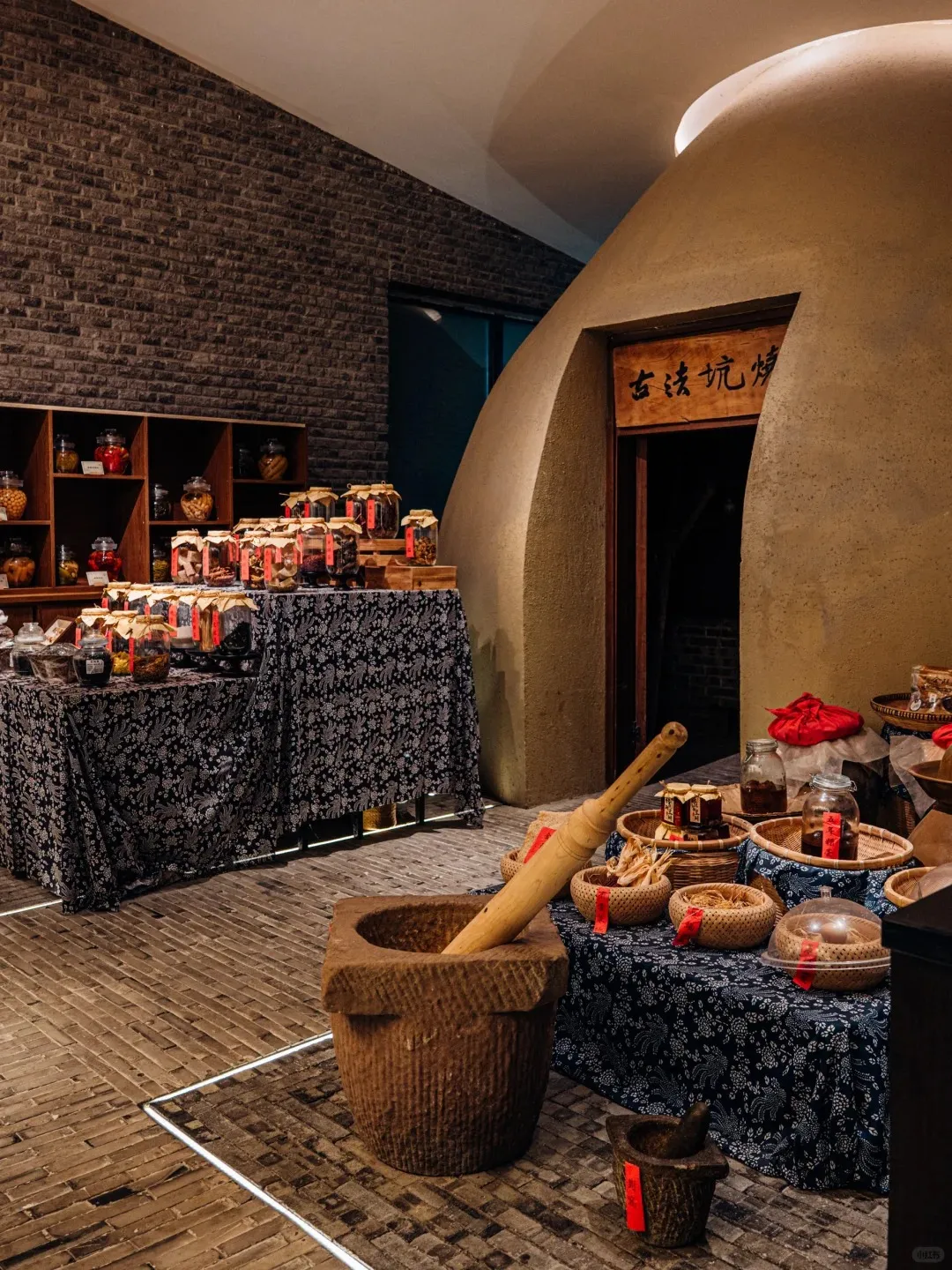 An indoor shop or exhibit showcasing traditional Chinese food products. On the left, a dark brick wall with wooden shelves holds jars of dried ingredients. In the center, tables draped in blue and white patterned fabric display numerous jars of preserved foods and woven baskets of dried herbs and spices. To the right, a large, dome-like, earth-toned structure with an arched entrance features a wooden sign reading '古法坑炖' (Ancient Method Pit Stew or Roasting). A large stone mortar with a wooden pestle sits prominently on the brick floor in the foreground, with a smaller mortar nearby.