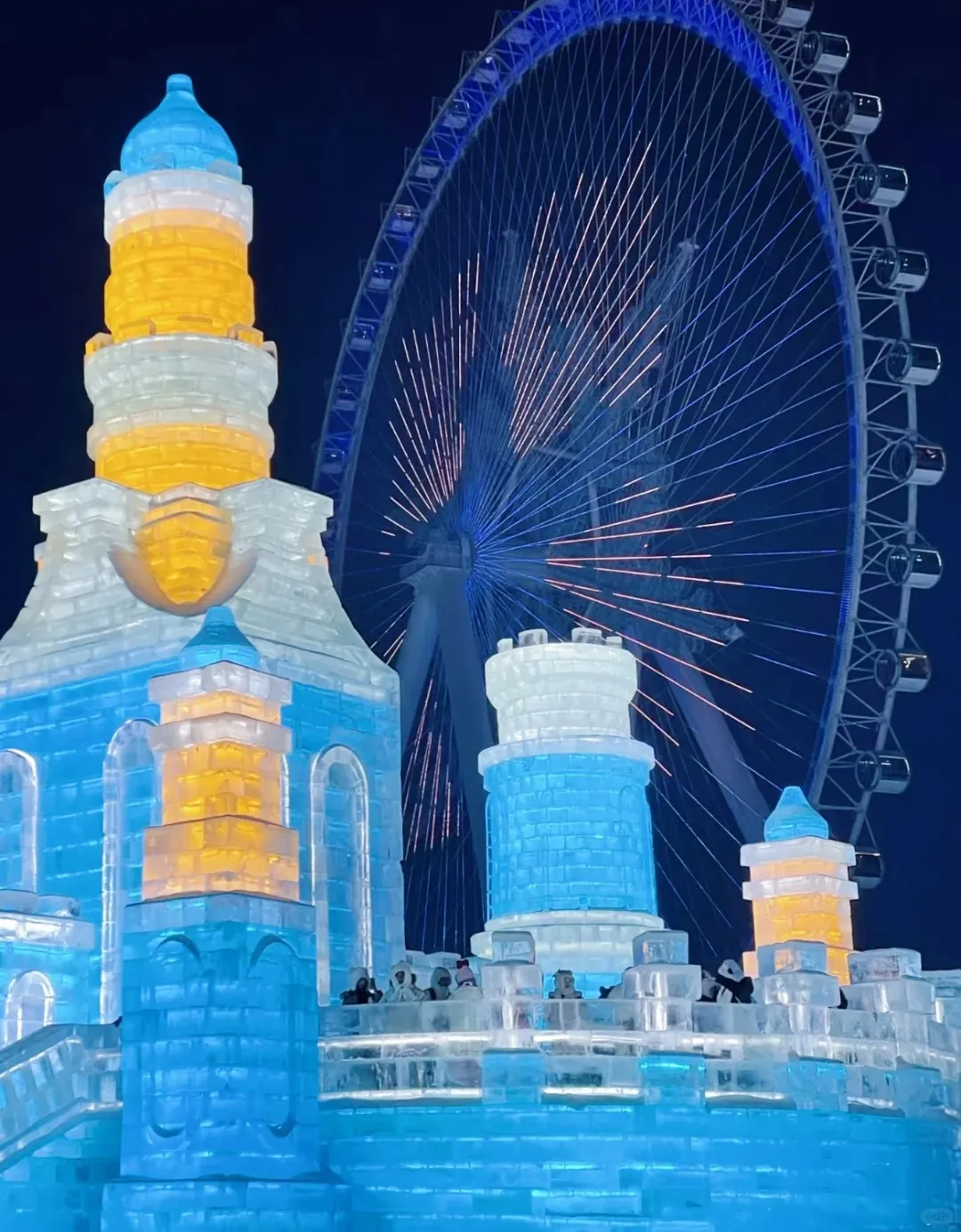 Nighttime image of the Harbin Ice and Snow Festival showing a large, illuminated ice castle with blue, white, and yellow sections. Several people are visible on the castle's lower platforms. In the background, a huge Ferris wheel glows with blue and pink lights against the dark sky.