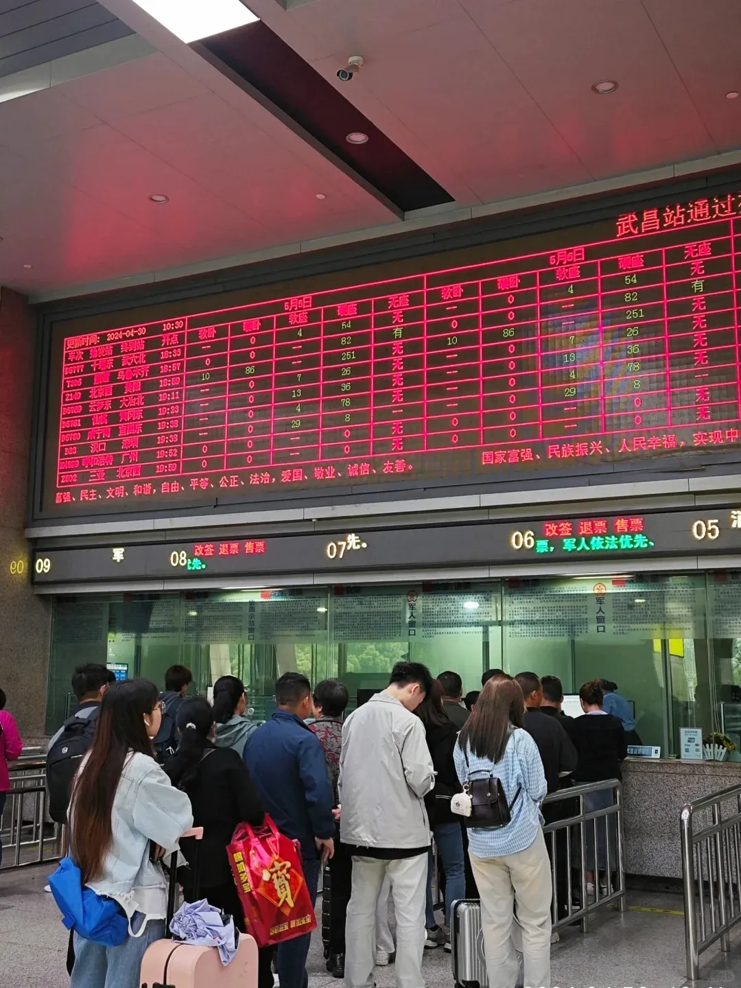 Travelers, some with luggage, queueing at manual ticket counters in a bustling Chinese high-speed railway station. A large red LED display board above shows real-time train schedules, destinations like Beijing and Guangzhou, departure times, and ticket availability. Passengers are waiting to book, change, or refund tickets, reflecting the process for international travelers using passports to navigate train travel in China.