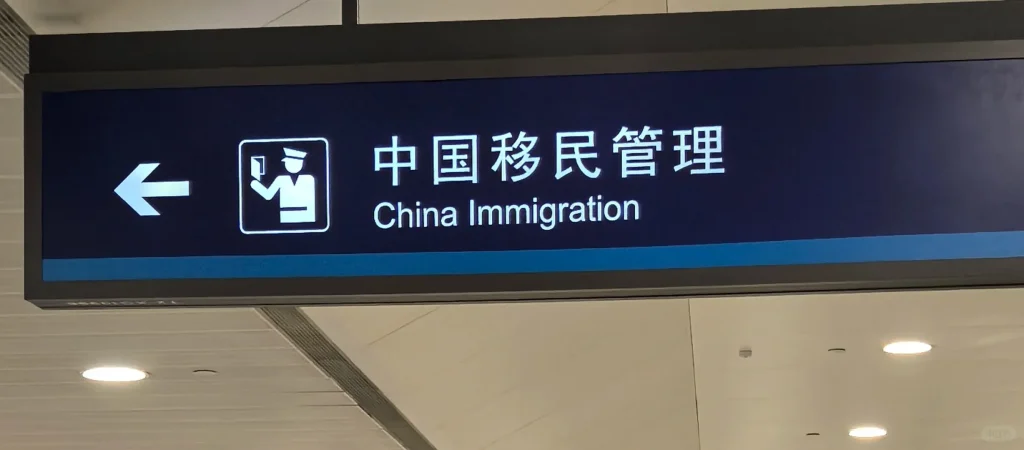 An illuminated airport sign with a dark blue background displaying '中国移民管理' and 'China Immigration' in white text. To the left is an icon of an officer holding a passport and a white arrow pointing left.
