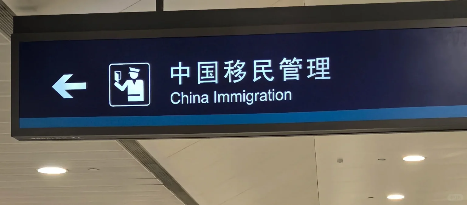An illuminated airport sign with a dark blue background displaying '中国移民管理' and 'China Immigration' in white text. To the left is an icon of an officer holding a passport and a white arrow pointing left.