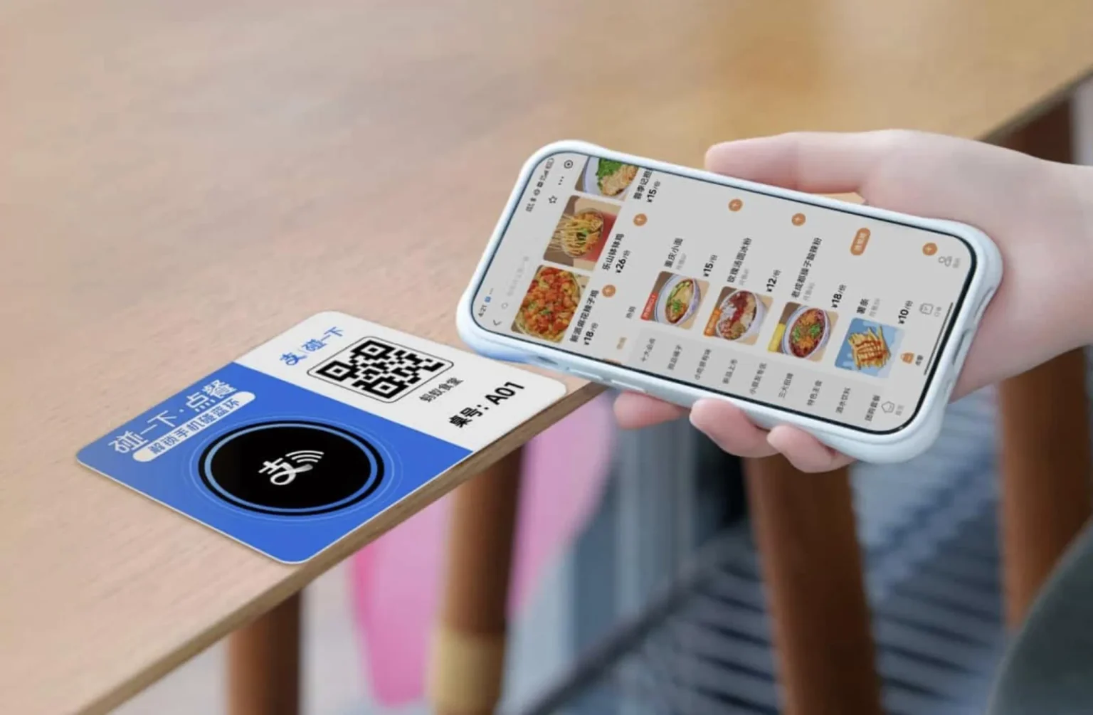 A person's hand holds a white smartphone over a blue card on a light wooden table. The smartphone screen displays a Chinese food ordering app with images of various dishes and prices. The blue card features a QR code, an NFC symbol, and Chinese text that translates to 'Touch to order' and 'Table number: A01'.