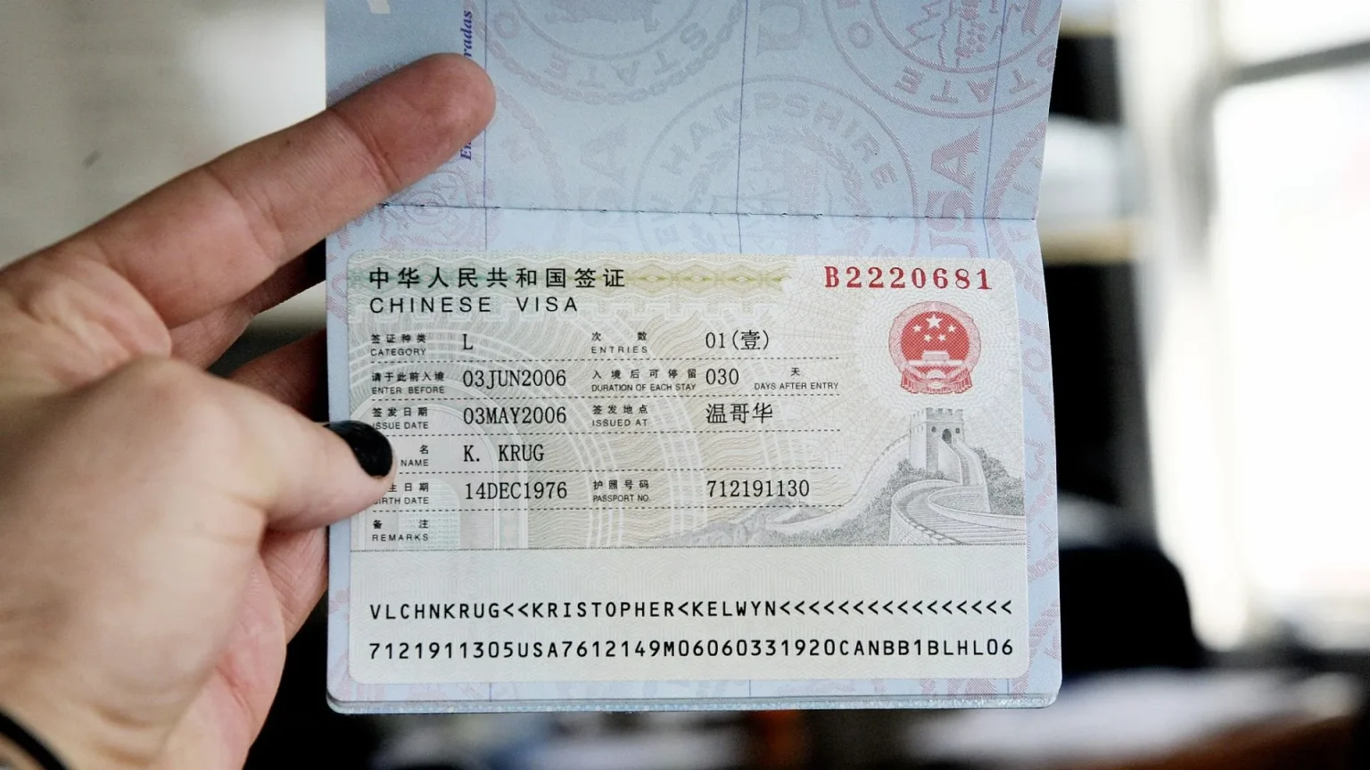 Close-up of a person's hand holding open a passport page, displaying a Chinese tourist visa sticker. The visa shows details like 'CHINESE VISA,' category 'L,' issue date '03MAY2006,' enter-before date '03JUN2006,' name 'K. KRUG,' passport number '712191130,' and features images of the Great Wall and the Chinese national emblem.