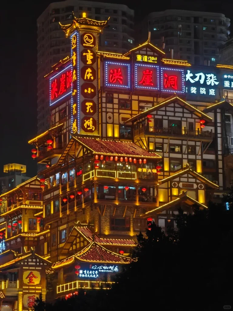 Multi-story traditional Chinese stilt house complex, identified as Hongya Cave by prominent red neon signs, brightly illuminated at night. Yellow string lights outline the building's tiered roofs, balconies, and wooden framework, where numerous red lanterns hang. Other yellow neon signs with Chinese characters are visible on a tall central tower. Modern high-rise buildings loom in the dark background.