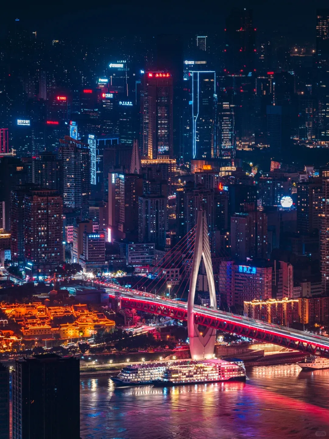 Panoramic night view of Chongqing city featuring a large illuminated ferry on the Yangtze River. The red-lit Chaotianmen Bridge, with its distinctive white arch pylon, spans the river carrying traffic. On the left riverbank, the multi-tiered, traditional-style Hongyadong complex glows with golden-orange lights. The background shows a dense skyline of modern high-rise buildings illuminated with colorful neon signs against the dark night sky.