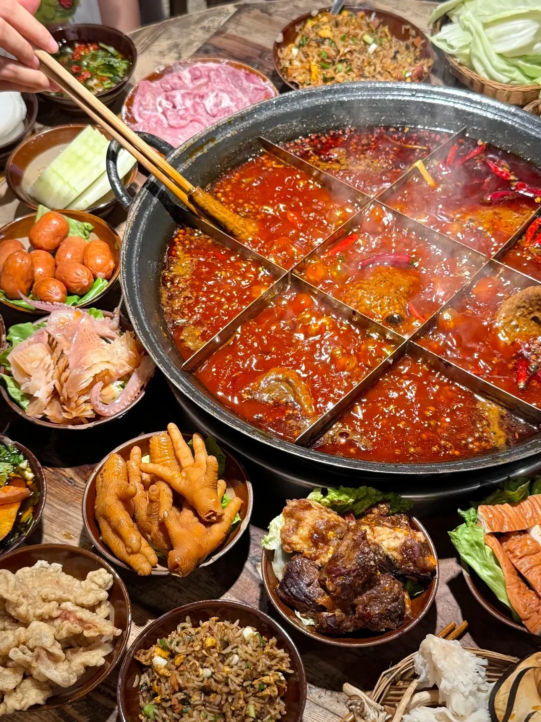 A large, black nine-grid Chongqing hotpot bubbling with red spicy broth, chilies, and Sichuan peppercorns, surrounded by an array of hotpot ingredients on a wooden table. These ingredients include thinly sliced raw beef, cooked chicken feet, crispy fried snacks, small sausages, sliced cucumber, fried rice, cabbage leaves, and various offal and fungi. A hand is seen using long chopsticks to place food into the pot.