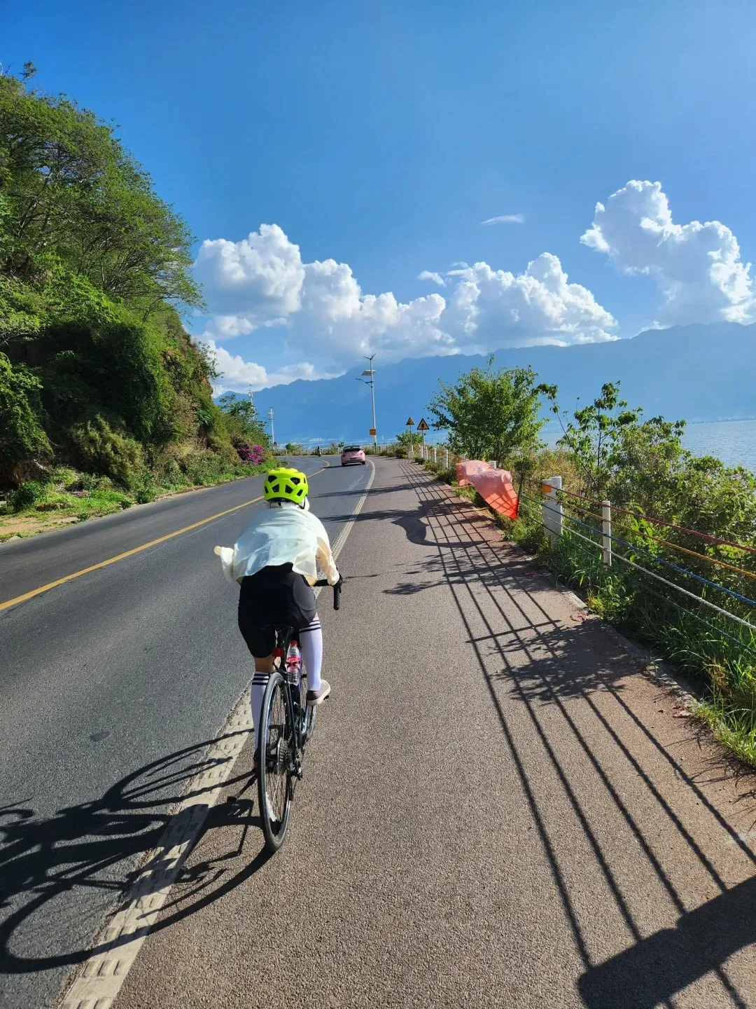 A person wearing a neon yellow helmet, white long-sleeve top, black shorts, and white knee-high socks cycles from behind along the paved ecological corridor of Erhai Lake in Dali, Yunnan. To the left, a main road runs parallel to a lush, green tree-covered hillside. To the right, the vast blue Erhai Lake stretches towards distant mountains under a bright blue sky dotted with fluffy white clouds. A pink car is visible in the distance on the main road, and strong shadows from the cyclist and a nearby fence indicate bright sunlight.