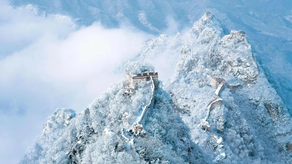 Snow-covered Great Wall of China winding across several rugged mountain peaks with ancient watchtowers, surrounded by snow-laden trees and misty clouds.