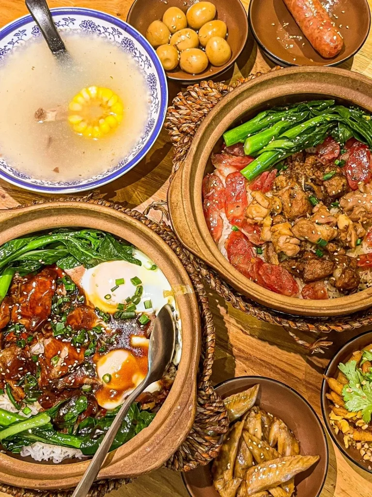 A vibrant spread of traditional Cantonese cuisine from Guangzhou, perfect for a meal beyond dim sum. The focus is on two steaming claypot rice dishes: one with sliced Chinese sausage (lap cheong), savory chicken, and fresh gai lan, and another with braised meat, two sunny-side-up fried eggs, and green vegetables over rice, all drizzled with sauce. The meal also includes a clear pork rib soup with a corn cob slice, a bowl of braised quail eggs, a separate whole Chinese sausage, glazed chicken wings, and a side of crispy fried snacks, all laid out on a rustic wooden table.