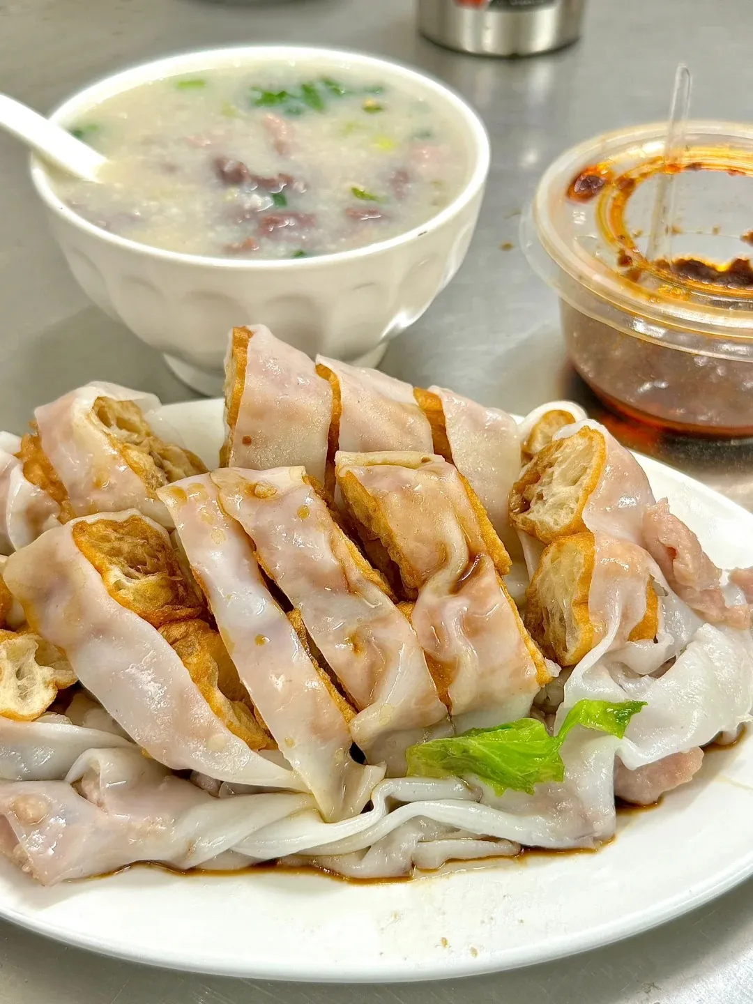 A traditional Cantonese breakfast meal featuring a plate of silky smooth rice noodle rolls (Chee Cheong Fun) with crispy fried dough (youtiao) filling, drizzled with soy sauce, next to a white bowl of savory congee (porridge) topped with meat and green onions, and a small container of chili oil, representative of Guangzhou food.