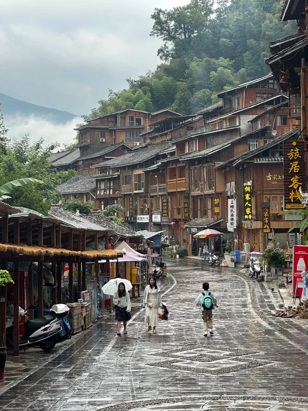 A traditional wooden stilt house village street on a rainy day. The wet cobblestone street, with decorative stone patterns, slopes uphill, lined with multi-story wooden houses featuring dark tiled roofs and balconies. Lush green trees and misty mountains are visible in the background. Several people are walking on the street, including two women with a white umbrella and a child with a blue backpack.