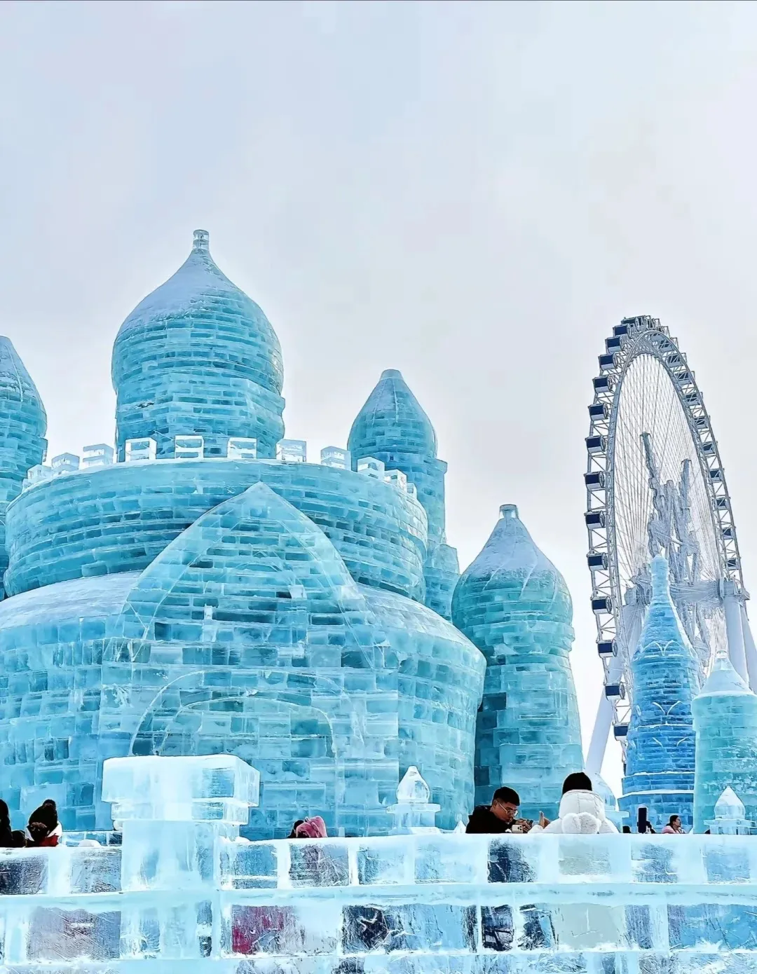 A massive, intricate castle structure built entirely from translucent blue ice blocks, featuring multiple domed towers and crenellated walls. A large white Ferris wheel is prominently visible in the background under a bright, overcast sky. Several visitors are gathered at the base of the ice sculpture.