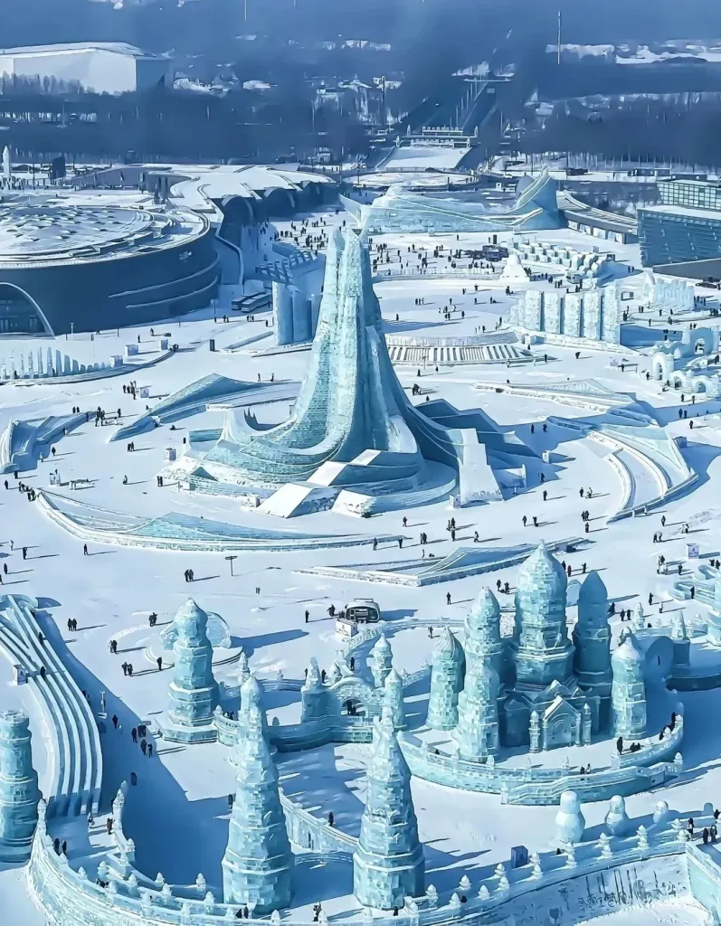 Aerial daytime overview of the Harbin International Ice and Snow Sculpture Festival, featuring a towering central ice spire, elaborate multi-spired ice castles, and vast icy pathways bustling with visitors at the Harbin Ice and Snow World.