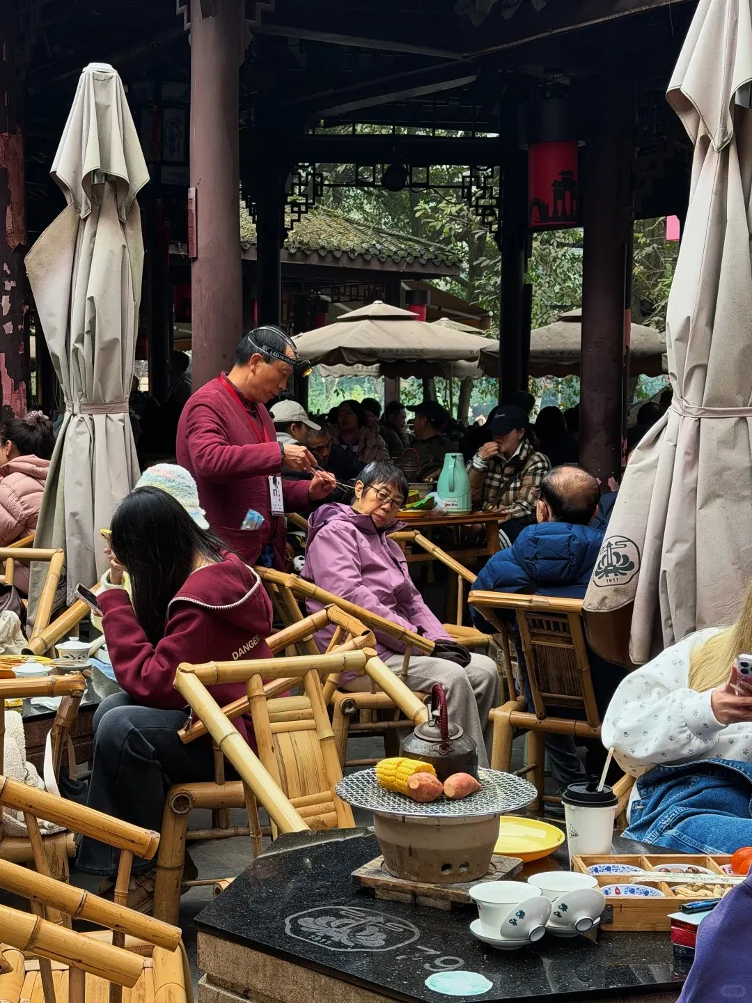 A bustling outdoor teahouse. In the foreground, a charcoal grill with corn and sweet potatoes sits on a black table with a dark teapot and white gaiwans. People sit on bamboo chairs. A man in a red jacket with a headlamp performs an ear cleaning service on a seated patron. Beige parasols and traditional wooden architecture are visible throughout the crowded scene.