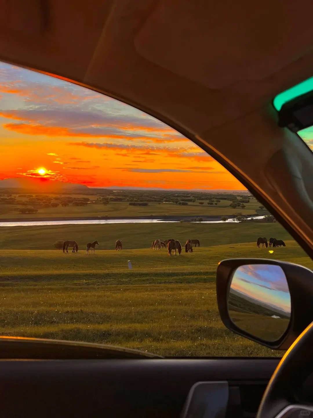 View from inside a car showing a vibrant orange and red sunset over expansive green grasslands. A herd of brown horses, including a foal, grazes near a winding river. The car's dark dashboard and a side mirror reflecting the sunset are visible in the foreground.