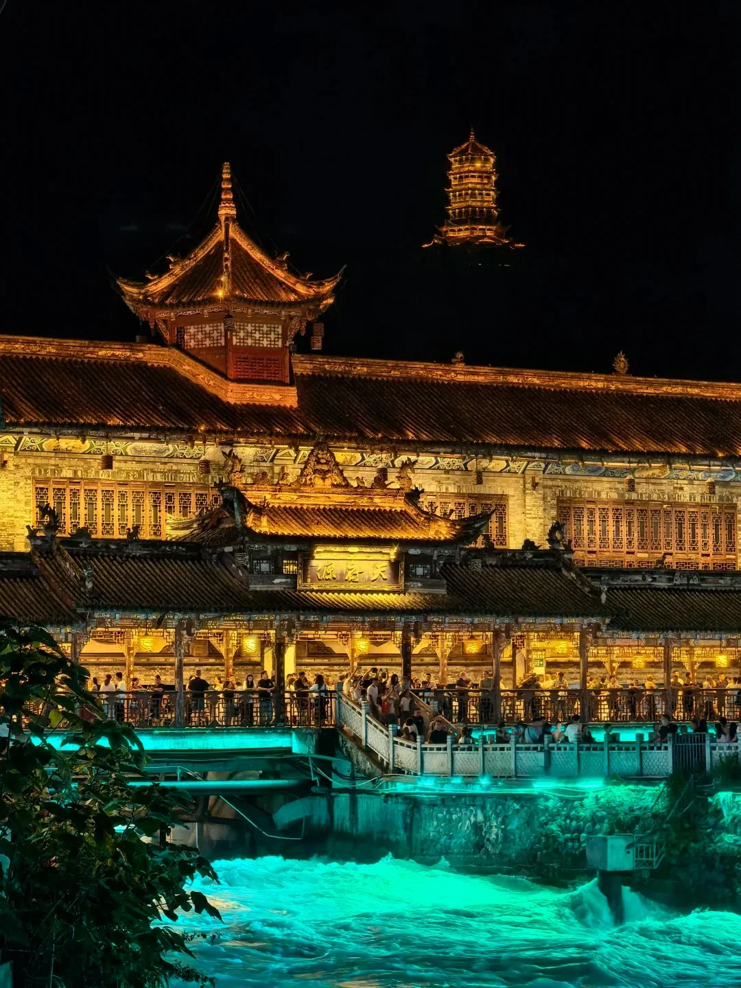 Lidui Park at Dujiangyan Irrigation System at night, showcasing traditional Chinese architecture brightly illuminated with warm golden lights. Below, the Min River rushes with a vibrant teal-blue glow from underwater lights, reflecting on the turbulent water. People gather on the park's balconies and a bridge that spans the river. In the upper background, a distant pagoda is also lit up, standing out against the dark sky, characteristic of this famous day trip from Chengdu.
