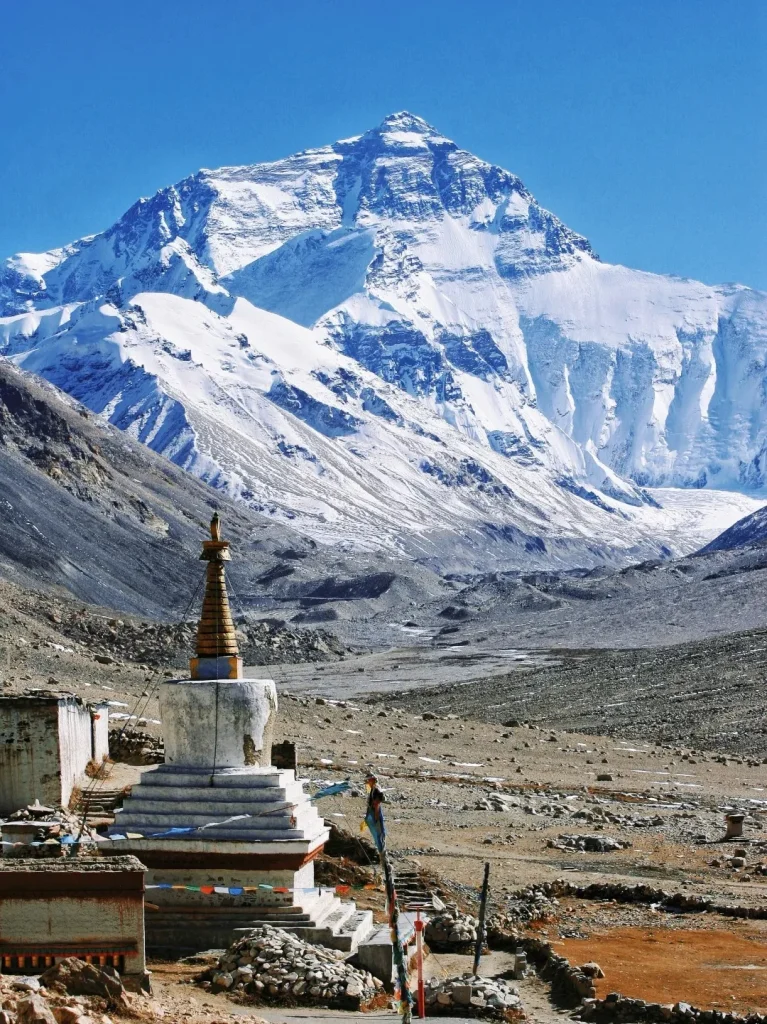 A breathtaking panoramic view of the majestic, snow-capped Mount Everest under a brilliant blue sky, as seen from Rongbuk Monastery in Tibet. In the foreground, a traditional white Buddhist stupa (chorten) with a golden spire and colorful prayer flags stands on a rocky, arid landscape. The path leads through a desolate valley towards the towering Himalayan peaks, highlighting this iconic Everest Base Camp view and its connection to required Tibet travel permits.