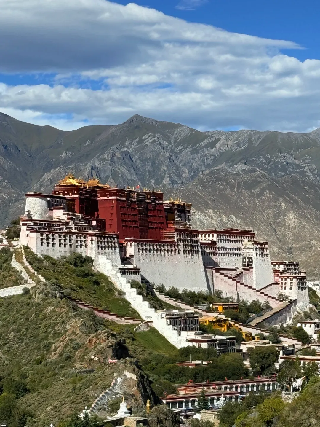 The Potala Palace in Lhasa, a grand multi-story complex featuring prominent red and white sections with golden roofs, stands majestically atop a green, rocky hill. Numerous white steps and terraced structures ascend the hillside towards the palace. In the background, extensive gray and green mountains stretch under a bright blue sky with scattered white clouds.