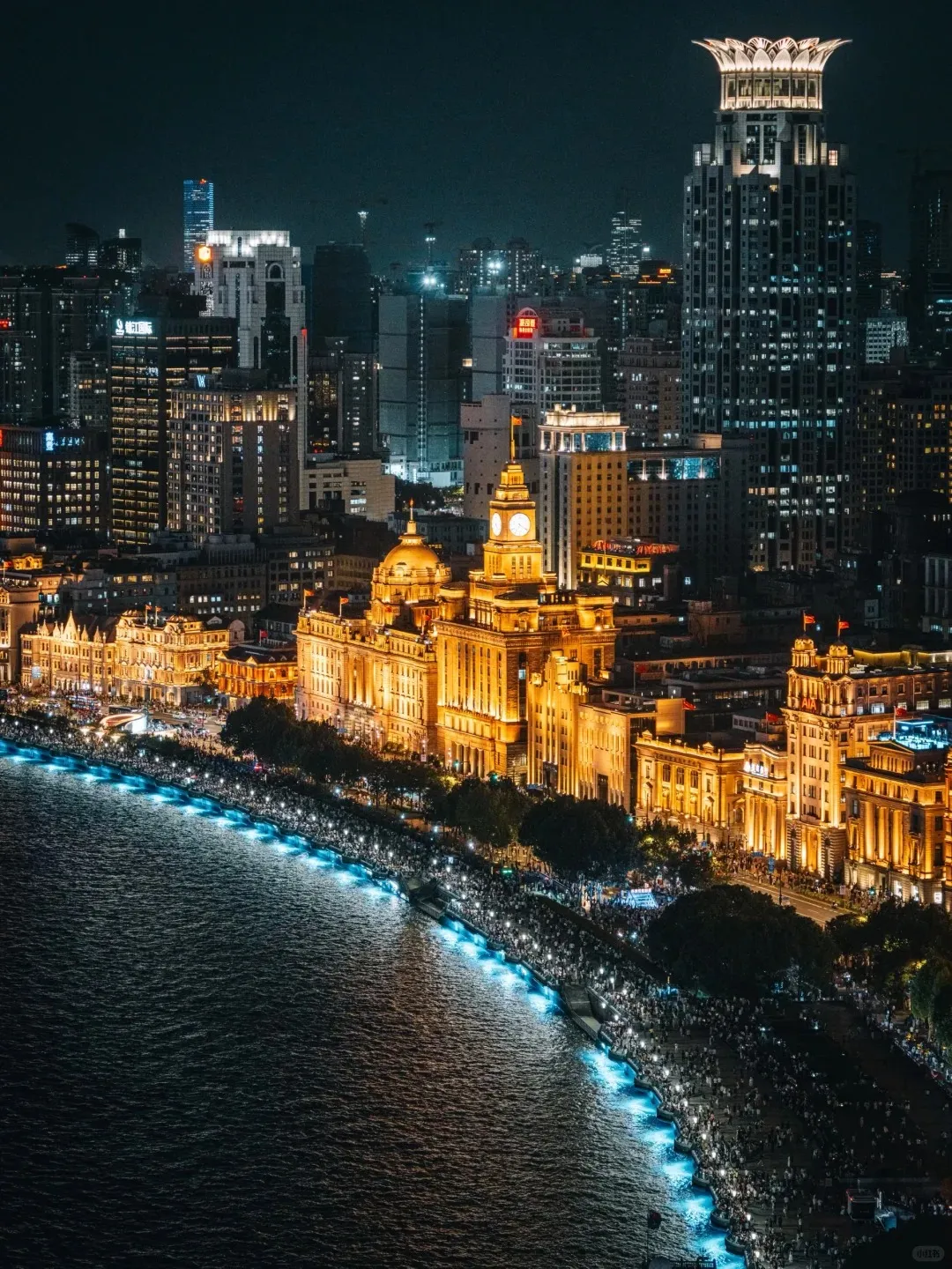 Panoramic aerial view of the Shanghai Bund at night. The Huangpu River runs along the bottom, beside a promenade illuminated by blue lights and filled with crowds of people. Behind the promenade, historic European-style buildings, including one with a prominent clock tower, glow with warm yellow light. In the background, numerous modern skyscrapers rise into the dark sky.