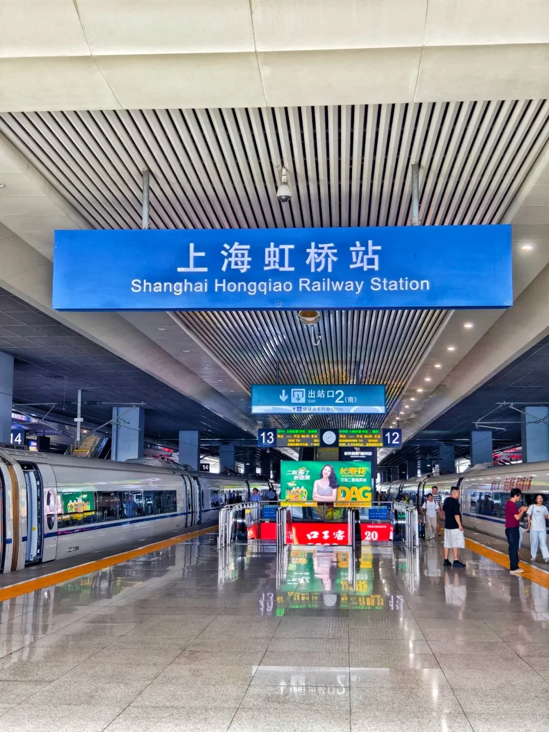 Interior view of Shanghai Hongqiao Railway Station platform with two white high-speed trains visible on tracks 14 and 12. A large blue overhead sign reads '上海虹桥站 Shanghai Hongqiao Railway Station'. The platform features red automated ticket gates, digital information screens, and several travelers. A smaller blue sign for 'Exit 2 (South)' is also visible.