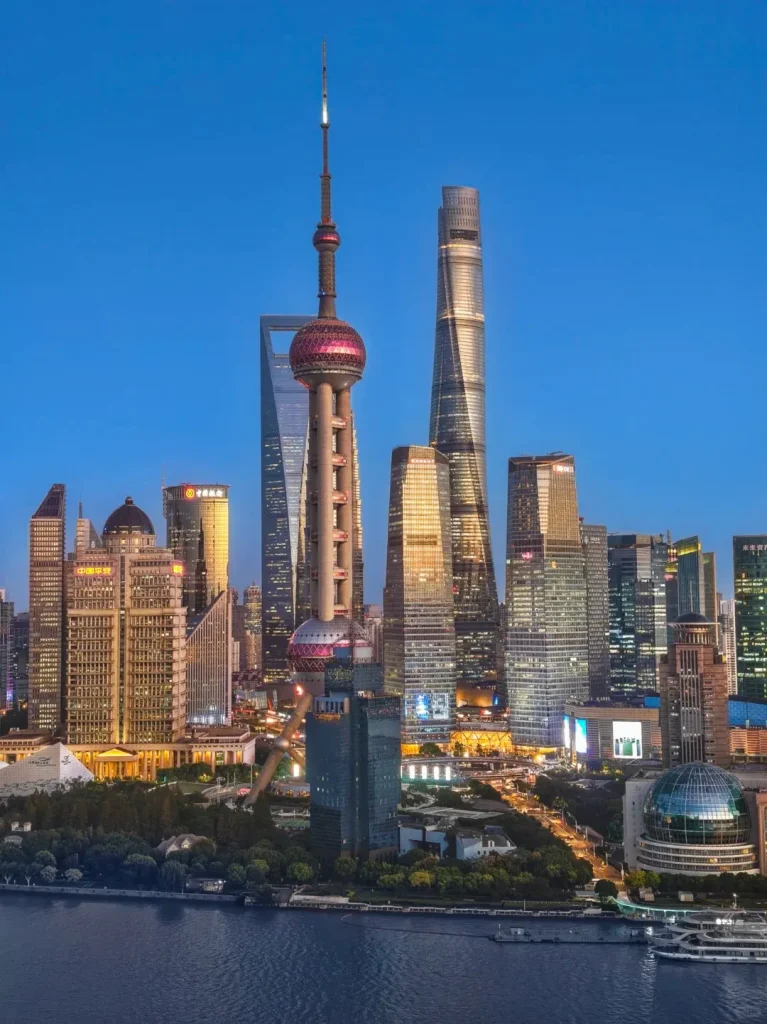 Panoramic view of the Shanghai Pudong skyline at twilight, featuring the illuminated Oriental Pearl TV Tower, Shanghai World Financial Center, and Shanghai Tower alongside numerous modern skyscrapers, with the Huangpu River in the foreground.