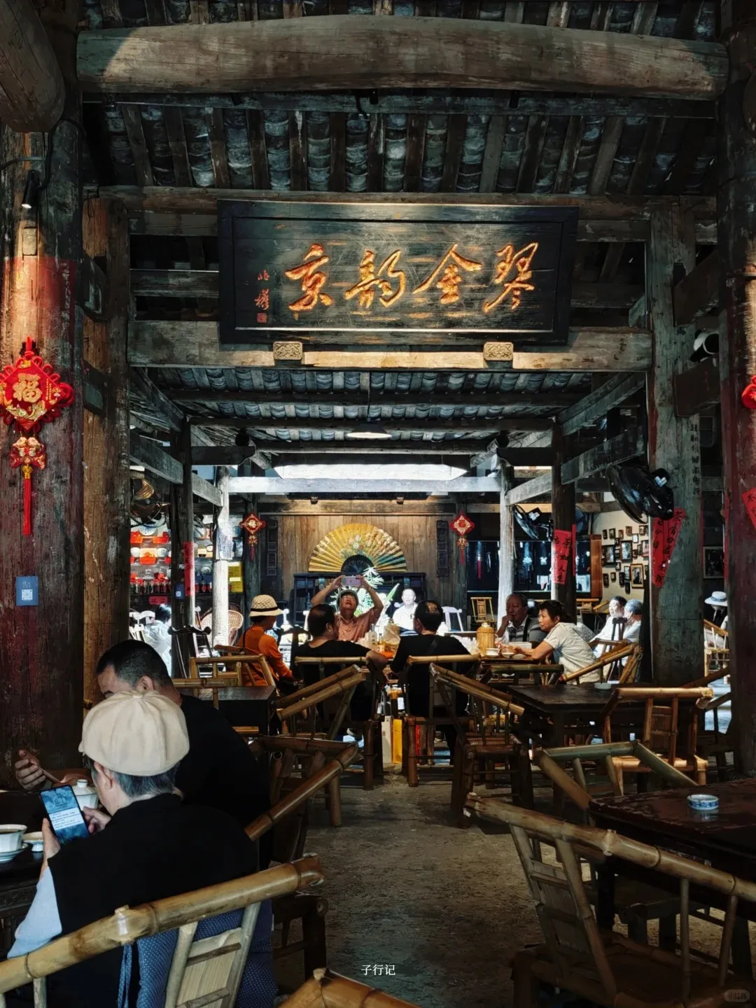 Interior view of a bustling traditional Chinese teahouse with high dark wooden beamed ceilings and a tiled roof structure. Numerous customers, mostly adults, are seated at dark wooden tables with light-colored bamboo chairs, drinking tea and socializing. A prominent rectangular wooden plaque with large gilded Chinese characters hangs centrally, with smaller red couplets on a wooden pillar to the left. Red festive decorations are visible on the pillars.