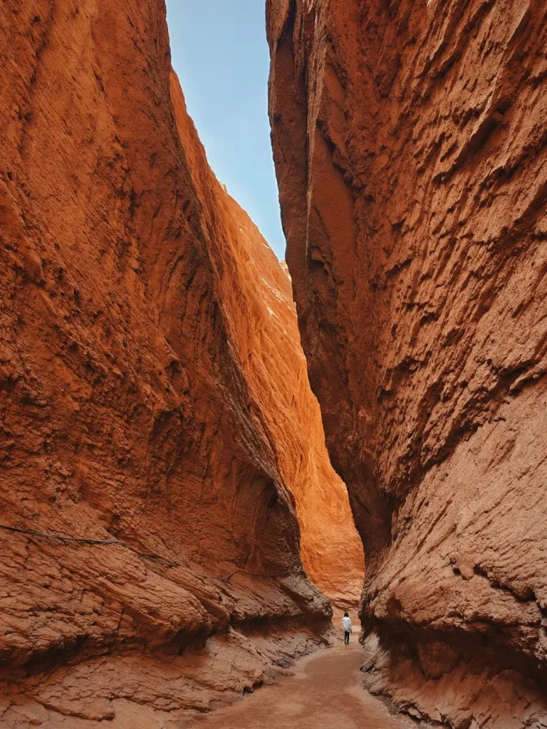 A lone person walks along a narrow, winding dirt path between towering, rugged red sandstone walls of the Tianshan Mystic Grand Canyon near Kuqa, Xinjiang, with a sliver of bright blue sky visible above.
