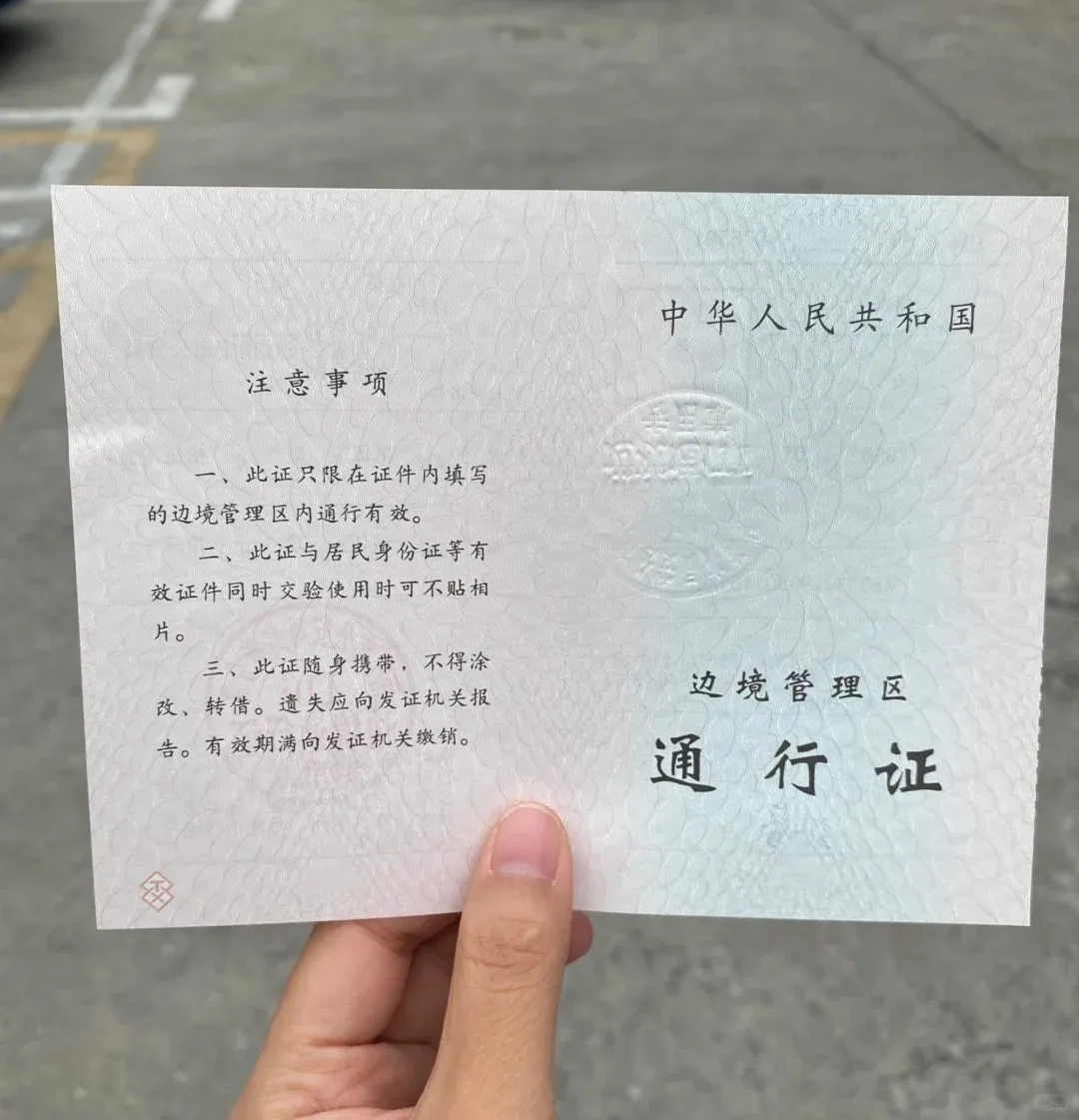 A close-up of a hand holding an official light blue-green 'Border Management Area Pass' (边境管理区 通行证) issued by the People's Republic of China. The document features an embossed seal in the center, the title '中华人民共和国' (People's Republic of China) at the top right, and '注意事项' (Notes/Precautions) on the left side, detailing its usage for specific border regions.