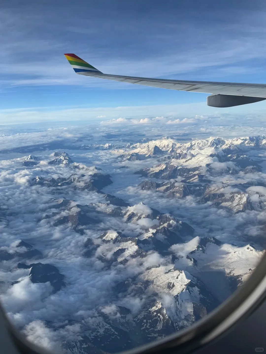 Aerial view from an airplane window, with the bottom right frame visible. A white airplane wing with a rainbow-colored wingtip extends into the upper left. Below the wing are vast snow-capped rocky mountains partially covered by white clouds under a clear blue sky.