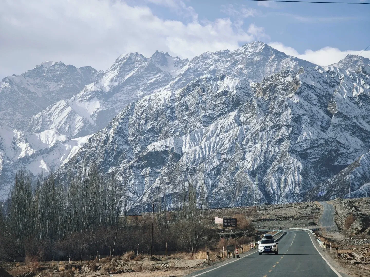 A paved road with a white SUV driving towards the viewer, winding through a valley flanked by bare trees and arid ground, with towering snow-covered mountains in the background under a partly cloudy sky.