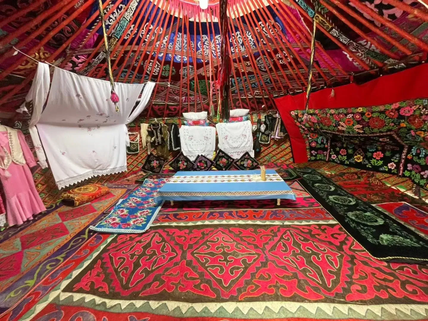 Interior of a traditional Mongolian yurt showcasing colorful patterned rugs on the floor, intricately embroidered textiles adorning the wooden lattice walls and red roof poles, and a low blue table in the center. A pink traditional garment hangs on the left.