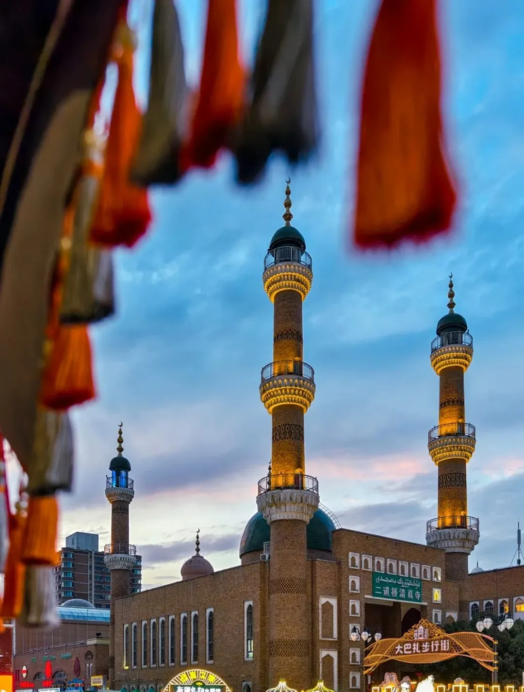 The grand Erdaoqiao Mosque at Urumqi Grand Bazaar in Xinjiang, illuminated brightly at twilight with its distinctive minarets and green domes, seen from below decorative red and white tassels hanging in the foreground, capturing the vibrant evening atmosphere.