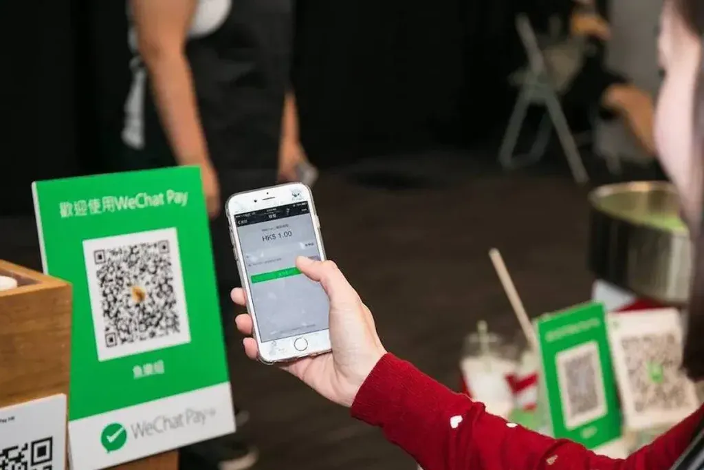 A close-up of a person's hand holding a white smartphone, scanning a QR code displayed on a green 'WeChat Pay' sign. The phone screen shows 'HKS 1.00' indicating a transaction in Hong Kong Dollars. Another smaller green QR code sign is visible in the blurry background.