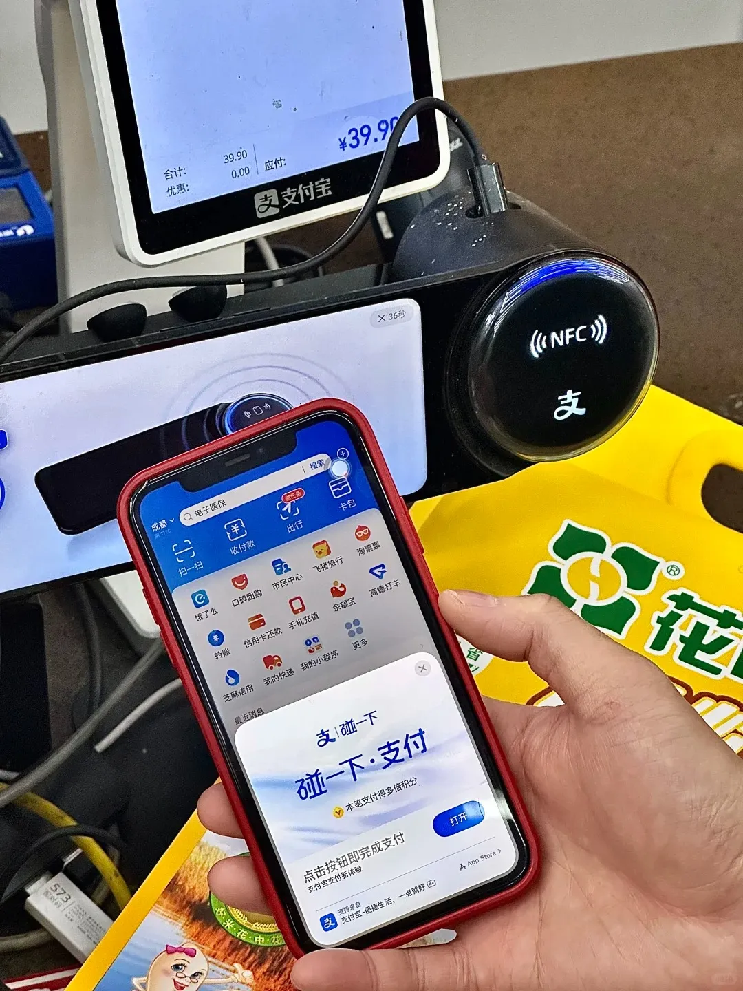 A person's hand holds a red-cased smartphone displaying the Alipay app's 'Tap to Pay' (碰一下·支付) screen. In the background, a payment terminal shows a total of ¥39.90 and an illuminated NFC reader with the Alipay logo.
