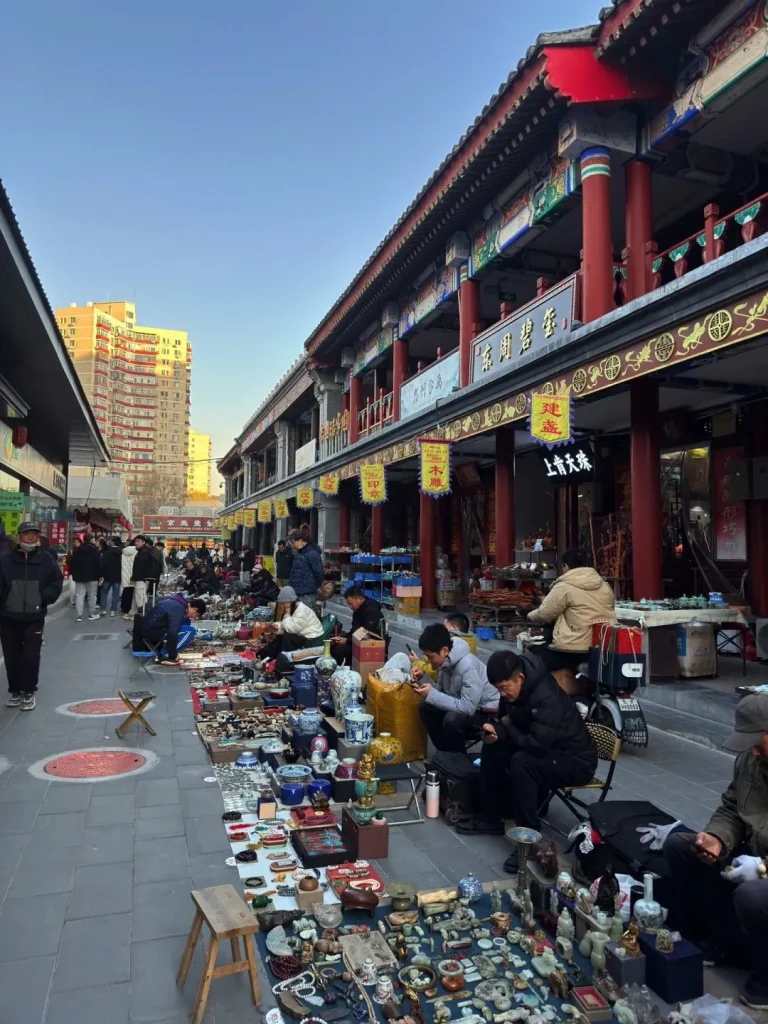 A bustling outdoor antique market in Beijing with numerous vendors displaying a wide array of curios, ceramics, jade figures, and other collectibles on cloths spread across the paved street. Many people are browsing or seated by the stalls. On the right, traditional Chinese buildings with red pillars and ornate roofs line the street, while modern high-rise residential buildings are visible in the distance under a clear blue sky.
