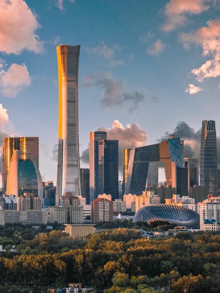 A vibrant sunrise or sunset over the modern Beijing cityscape, featuring prominent skyscrapers like the tall, slender CITIC Tower (China Zun) reflecting golden light, the distinctive CCTV Headquarters building, and the spiraling dome of the Phoenix International Media Center. A dense canopy of green and golden trees forms the foreground under a sky with dramatic, colorful clouds.