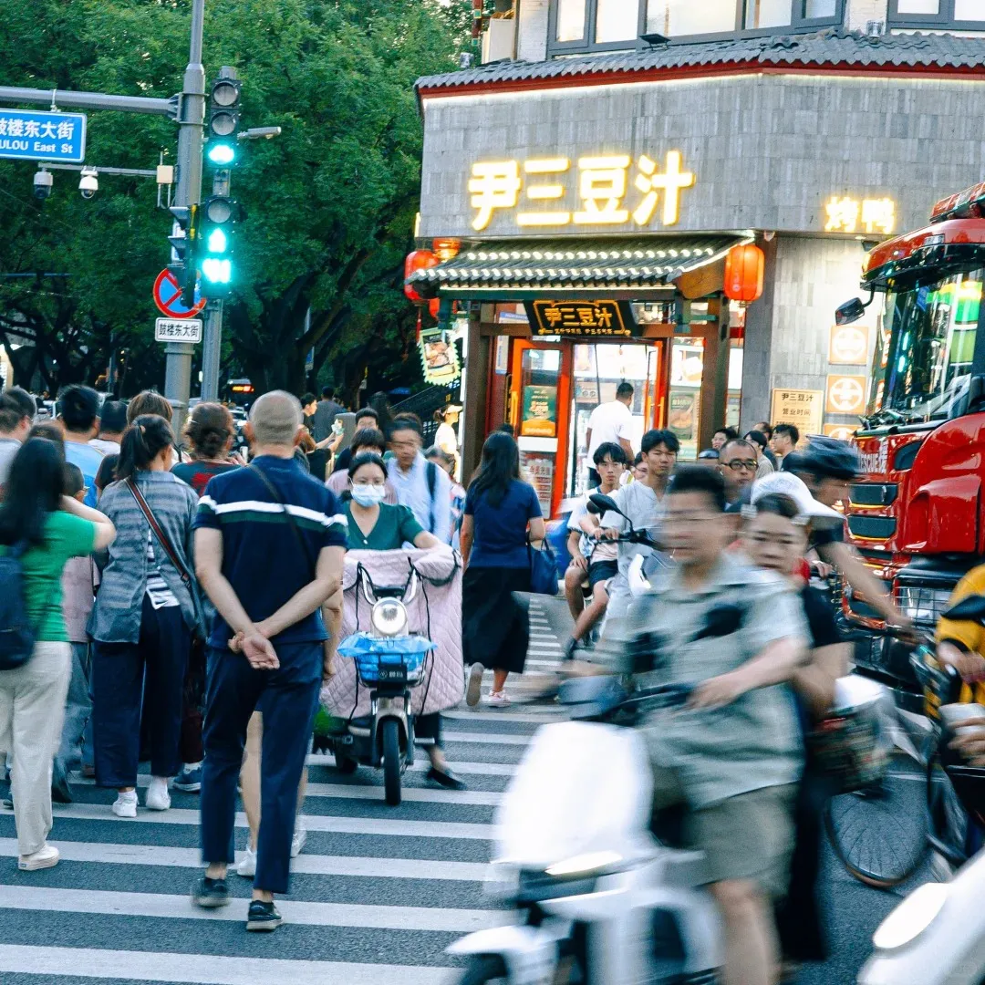 A bustling street in Beijing showing a large crowd of pedestrians and many electric scooters on a zebra crossing. A green traffic light is visible, and in the background, a building features glowing yellow Chinese characters on its sign. A blue street sign identifies the location as 'Gulou East St'.
