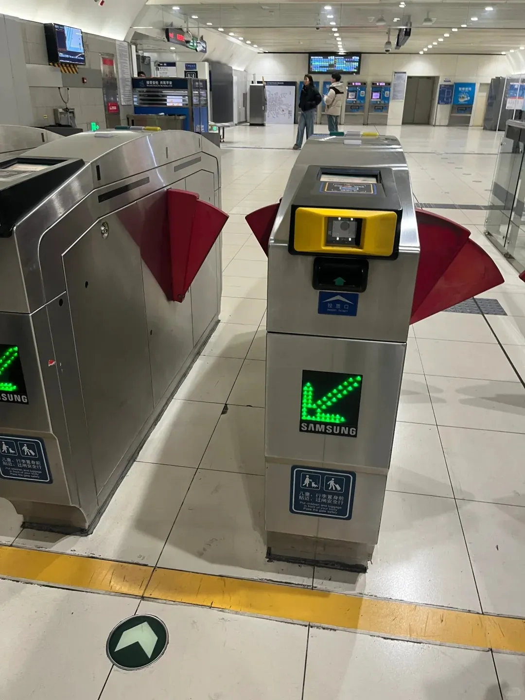 Two stainless steel subway turnstiles with red gates stand on a tiled floor in a Beijing metro station. Each turnstile displays a green LED arrow, a Samsung logo, and a blue safety sign with pictograms and text in Chinese and English. The turnstile on the right also has a yellow fare scanner.