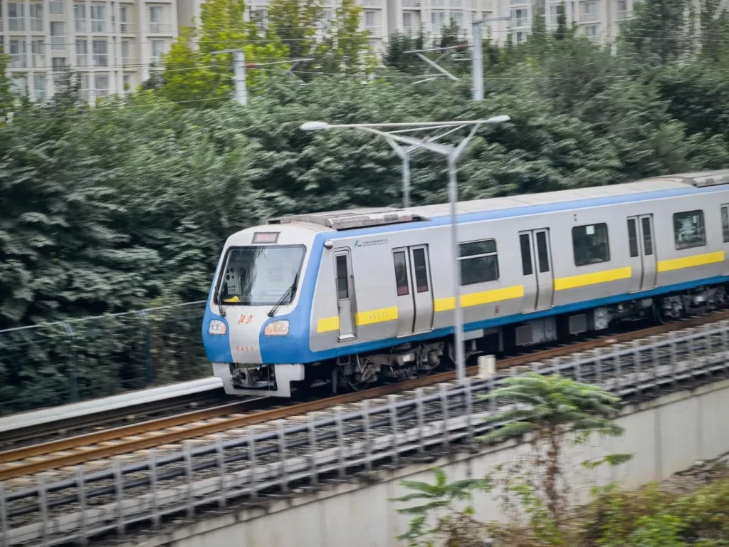 A modern Beijing subway train, silver with a blue front and a yellow stripe along its side, travels at speed on elevated tracks. Blurred green trees and tall white apartment buildings are visible in the background.