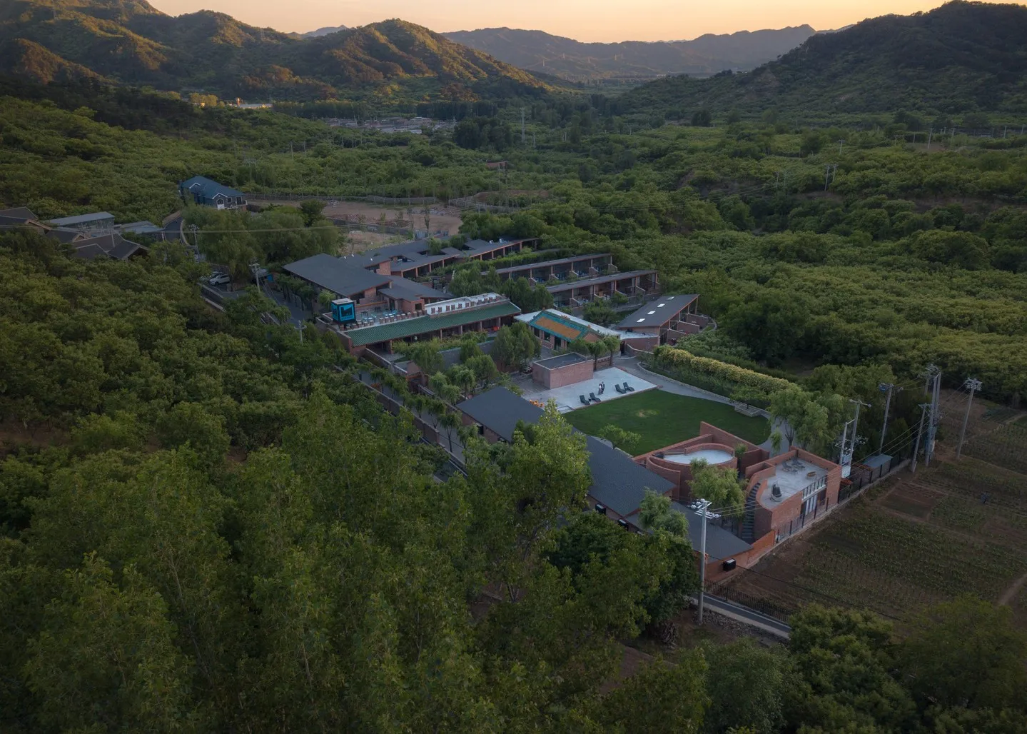 Aerial view of a modern resort complex with dark roofs and red brick walls, nestled within dense green forests and surrounded by mountains under an orange sky, with cultivated fields to the right.