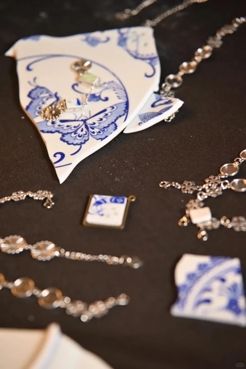 Jewelry made from upcycled blue and white porcelain shards, featuring a large butterfly-patterned pendant on a silver chain, and smaller rectangular and square porcelain pieces set in metal frames, alongside various silver jewelry findings, all laid on a dark surface.