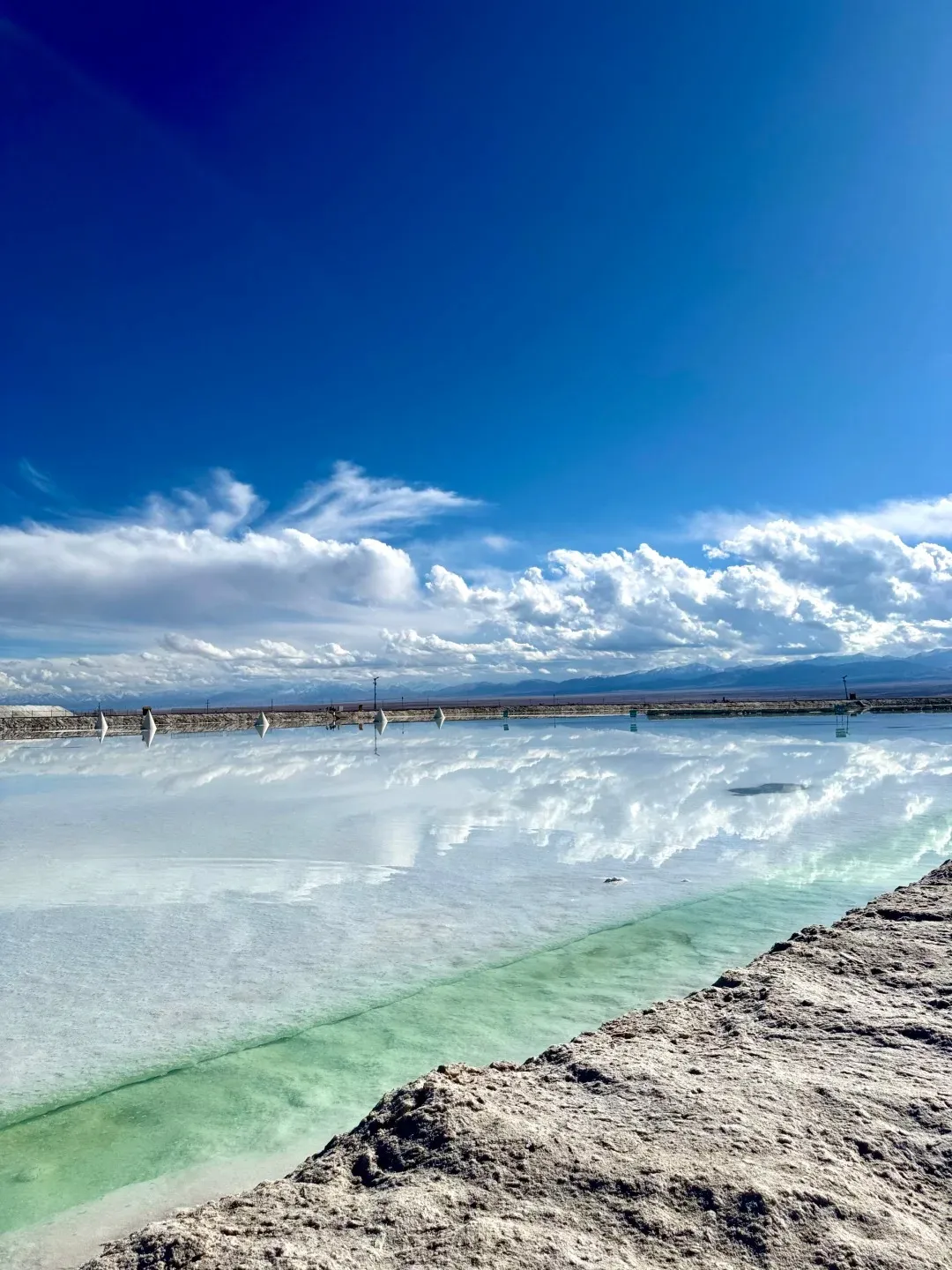 A panoramic view of Chaka Salt Lake, showing a vibrant blue sky with fluffy white clouds reflected on the lake's calm, pale blue and green surface. A textured, salt-encrusted bank occupies the foreground, sloping from the lower right, and a distant mountain range is visible beneath the horizon.