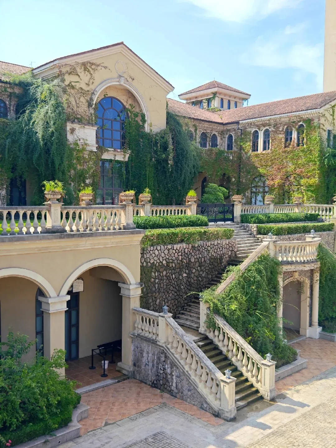 Exterior view of the Chengdu Contemporary Image Museum, a large building featuring light yellow walls, a terracotta tiled roof, and numerous arched windows, including a prominent large arched window. Lush green vines heavily adorn the facade. A grand stone staircase with white balustrades descends to a lower tiled courtyard, bordered by stone walls and manicured hedges.